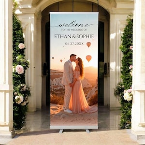May include: A wedding banner with the text "welcome to the wedding of Ethan & Sophie 06.27.20XX". The banner features a couple kissing with hot air balloons in the background. The banner is displayed on a white stand.