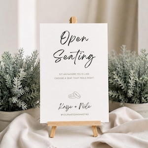 May include: A white sign on a small wooden easel with the words "Open Seating" in elegant black script. The sign includes the text "Sit anywhere you'd like, choose a seat that feels right," and the names "Kaye + Nile" with a wedding hashtag.