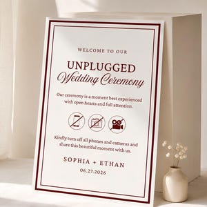 May include: A rectangular wedding sign with a white background and dark red border. The text reads: WELCOME TO OUR UNPLUGGED WEDDING CEREMONY. Our ceremony is a moment best experienced with open hearts and full attention. Icons show a phone, camera, and video camera with a slash through them. Text below: Kindly turn off all phones and cameras and share this beautiful moment with us. SOPHIA + ETHAN 06.27.2026.