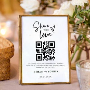 Op de afbeelding: Een goudkleurig ingelijst bord met de tekst "Share the Love" en een QR-code. Het bord bevat ook de namen "Ethan en Sophia" en de datum "06.27.20XX". De achtergrond is wazig met bloemen en kaarsen.