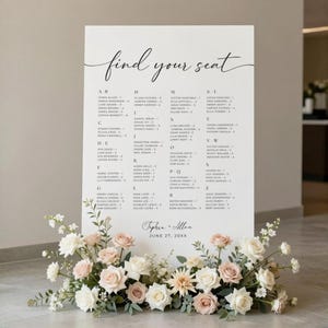 May include: A large white wedding seating chart with elegant calligraphy that reads "find your seat." The chart lists names alphabetically. A floral arrangement of white and blush roses and greenery sits at the base. The date is June 27, 20XX.