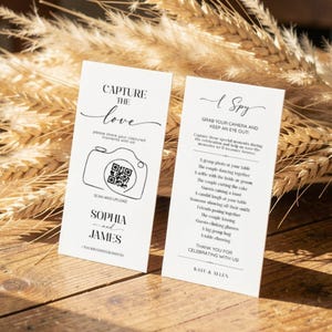 May include: Two white rectangular wedding cards with black text and a camera graphic. One card reads "Capture the Love" and includes a QR code. The other card says "I Spy" and lists prompts for guests. Both cards are on a wooden surface.