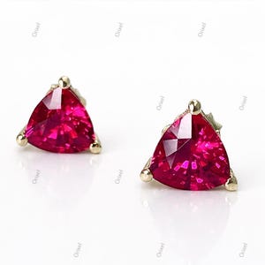 May include: A pair of gold stud earrings, each featuring a triangular, faceted, deep red gemstone. The earrings have a simple design with small gold accents at each corner, set against a white background.