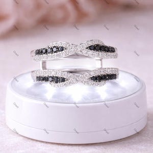 May include: Two silver rings with an intertwined design. Each ring features a row of black stones in the center, surrounded by smaller white stones. The rings are displayed on a white, illuminated platform, showcasing their intricate details.