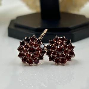 May include: Rose gold earrings, each with a cluster of deep red gemstones. The earrings are designed in a floral pattern, with each gemstone meticulously set to create a sparkling effect. The earrings are displayed on a white surface.