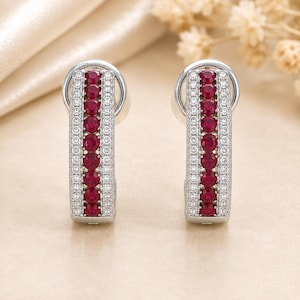 May include: A pair of silver-toned hoop earrings featuring a central vertical row of round, deep red stones flanked by two parallel rows of small, clear, sparkling stones. The earrings have a polished metallic finish and a secure hinged closure.