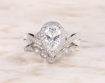 May include: A silver-toned engagement ring set featuring a pear-shaped center stone surrounded by a halo of small round stones. The band has a crisscross design encrusted with pavé stones, paired with a curved wedding band with round stones.