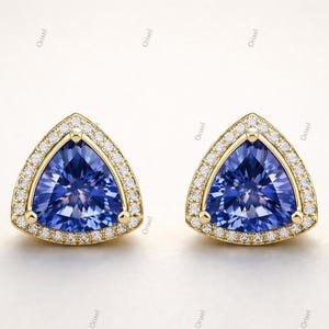 May include: A pair of gold stud earrings with a triangular, deep blue gemstone center. The gemstones are surrounded by a halo of small, clear diamonds. The earrings are set against a white background.