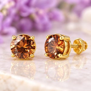 May include: A pair of stud earrings featuring round-cut, deep amber-colored gemstones set in polished yellow gold. Each earring is secured with a threaded screw-back post, showcasing a faceted design that reflects light across the warm brown surface.