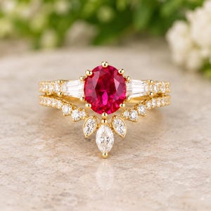 May include: A gold ring set featuring a central oval-cut vibrant red gemstone flanked by two baguette-cut clear stones. The band is adorned with small round clear stones, and a matching chevron-shaped guard ring features marquise-cut stones.