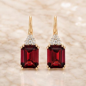 May include: A pair of gold drop earrings featuring emerald-cut deep red gemstones. Each earring is accented with a cluster of small, clear, round-cut stones at the top, set in a gold frame with a secure lever-back closure mechanism.