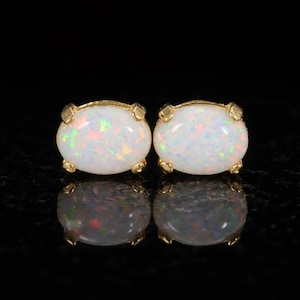 May include: A pair of oval-shaped opal stud earrings. The opals are white with iridescent flashes of color. Each opal is set in a gold-colored bezel with four prongs. The earrings are displayed on a reflective black surface.