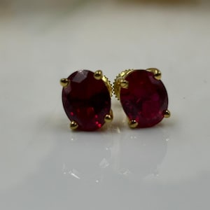 May include: A pair of gold stud earrings with oval-shaped, deep red gemstones. The earrings have a classic design, with the gemstones held in place by four small prongs. The earrings are set against a white background.