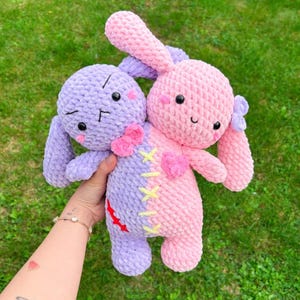 Pode incluir: Dois coelhos de crochê feitos à mão em tons de roxo e rosa. Os coelhos estão unidos, com um lado roxo e o outro rosa. Eles têm olhos pretos, bochechas rosadas e laços. Os coelhos estão em um fundo gramado verde.