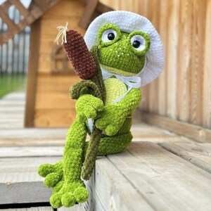 May include: A handmade green crochet frog wearing a white hat and holding a brown cattail. The frog has large, expressive eyes and is sitting on a wooden surface. The frog is a whimsical, decorative item.