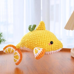 May include: A handmade crochet fish toy in bright yellow, orange, and white. The fish has a fin on its back and a green leaf accent. The fins and tail are designed to look like citrus fruit slices. The toy has a cute, friendly expression.