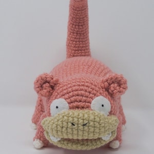 May include: A handmade crochet plush toy in the shape of a Slowpoke character. The plush is primarily pink with a light tan mouth and white accents on the eyes and feet. The tail is long and upright, with a pink tip. The toy is made of yarn.