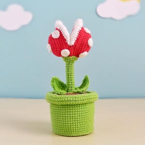 May include: A handmade crochet Piranha Plant from the Super Mario video game series. The plant features a red head with white spots, white teeth, and green leaves, all in a green pot. The plant is a whimsical, decorative item.
