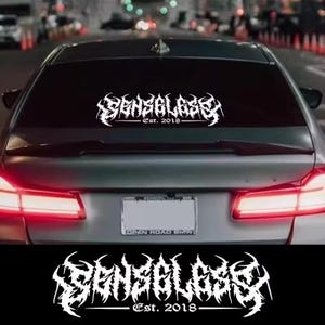 May include: White vinyl car decal with the word "SENSEGLASS" in a stylized font, with "Est. 2018" below. The decal is applied to the rear window of a silver car, with blurred city lights in the background.