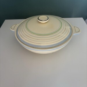 May include: A round, cream-colored ceramic casserole dish with a matching lid. The dish features horizontal pinstripes in shades of light blue and sage green circling the body and lid. It includes two side handles and a central knob on the lid.