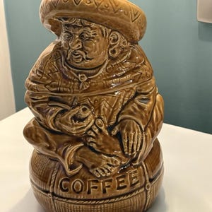 May include: A brown ceramic coffee jar in the form of a seated man wearing a sombrero. The jar features a detailed, textured surface, with the word "COFFEE" embossed on the base. The figure's clothing and hat are intricately designed.
