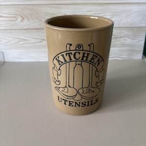 May include: A cylindrical tan ceramic utensil holder featuring a black graphic print of two ladles and a central tool, enclosed by a banner with the text KITCHEN UTENSILS. The container has a smooth, glossy finish and a wide, open top rim.