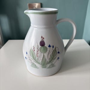May include: A white ceramic jug featuring a painted botanical design of a purple thistle, green leaves, and small blue flowers. The jug has a curved handle, a wide base, and a green painted rim around the top opening of the vessel.