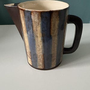 May include: A ceramic pitcher featuring a vertical striped pattern in shades of dark blue and speckled beige. The vessel has a solid dark brown handle and a matching dark brown triangular spout, with a smooth cream-colored interior finish.