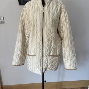 Puede incluir: Una chaqueta acolchada de rayas color crema y beige con cuello alto, cremallera frontal completa y dos bolsillos de parche con ribete beige sólido. La prenda tiene mangas largas y un patrón de costuras en forma de diamante en toda la tela.