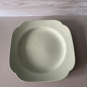 May include: A square-shaped ceramic plate with rounded corners and a smooth, pale mint green finish. The plate features a wide, flat rim that transitions into a shallow, circular centre basin, presenting a clean and uniform solid colour surface.