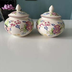 May include: Two matching cream-coloured ceramic jars with lids. Each jar features a floral pattern in pink, blue, yellow, and green. The lids have a red trim and a small, white knob. The jars are round and sit on a white surface, ideal for storage.