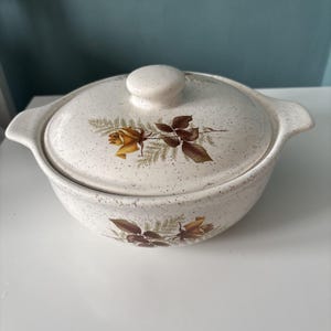 May include: A cream-colored ceramic casserole dish with a matching lid, featuring a speckled finish and a botanical print of yellow roses and green fern leaves. The round vessel includes two side handles and a central knob on the lid.