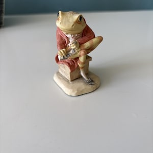 May include: A figurine of a frog dressed in a red coat and striped pants, seated on a small bench. The frog has a light tan complexion and is holding a small object. The base of the figurine is off-white.