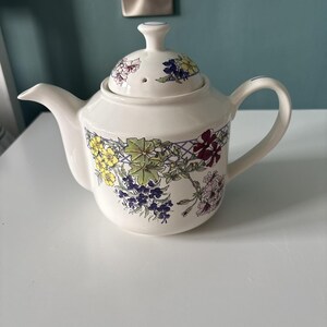 以下が含まれることがあります： 黄色、青、緑、濃い赤の花と葉のカラフルな植物柄が描かれた白い陶磁器のティーポット。ティーポットには、小さなつまみが付いたお揃いの蓋、湾曲したハンドル、注ぎ口があり、すべて花柄のモチーフで装飾されています。