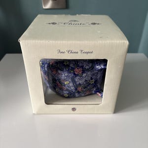 May include: A fine china teapot inside a white cardboard box. The box features the text "Chintz" and "Fine China Teapot". The teapot is visible through a rectangular cutout and displays a blue floral pattern with pink and yellow flowers.
