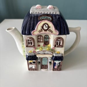 Può includere: Una teiera in ceramica a forma di edificio di un negozio di antiquariato a due piani. La struttura presenta un tetto blu scuro, pareti color crema, un orologio e la parola ANTIQUES stampata sulla facciata. Include beccuccio e manico bianchi.