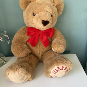 May include: A plush tan teddy bear with a dark brown nose and a bright red velvet bow around its neck. The bear has light tan paw pads, with the word FRASER embroidered in red thread on the left foot pad, featuring soft fur texture.