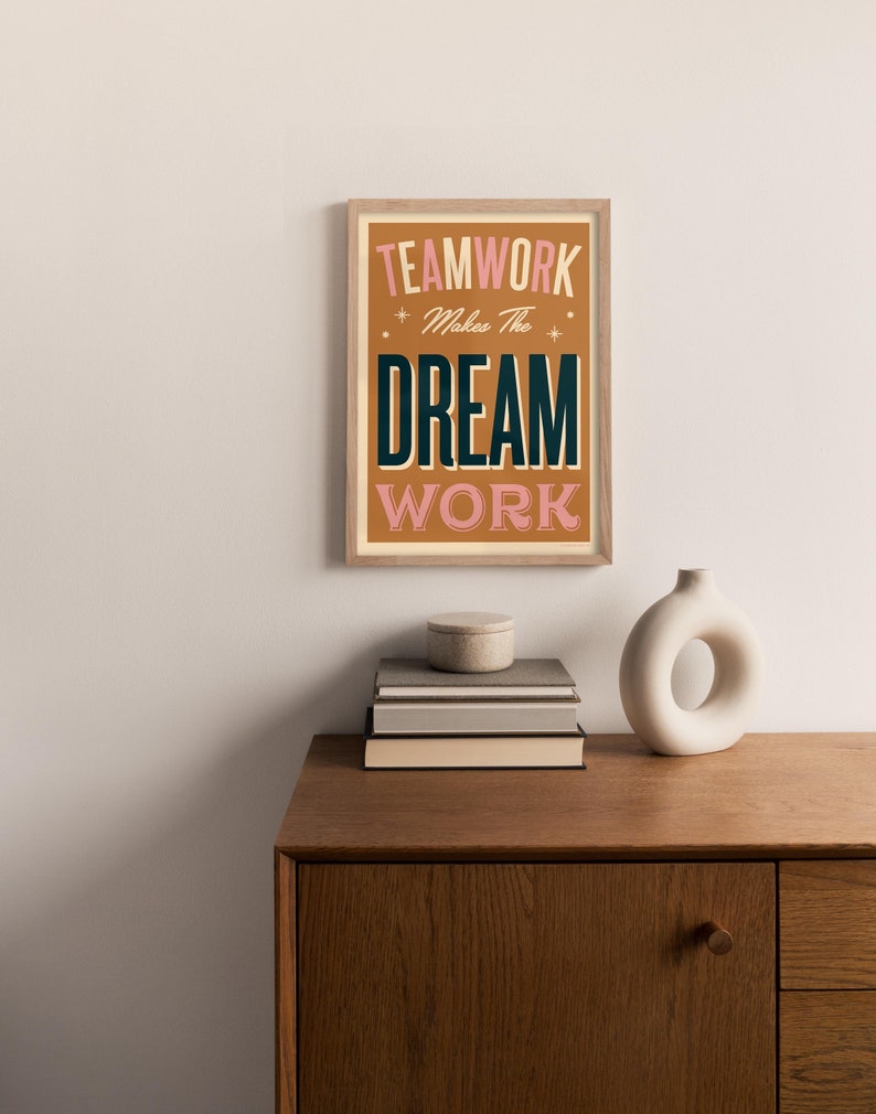 May include: A framed print with a motivational quote in a retro style. The quote reads "Teamwork Makes The Dream Work" with the words "Teamwork" and "Work" in black and the words "Makes The" and "Dream" in pink.