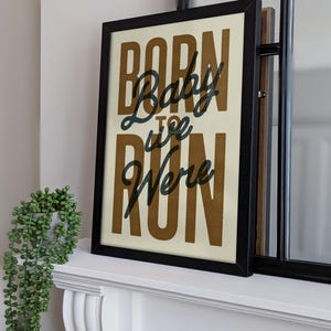 May include: A framed print with the text "Born to Run" in a brown font with the word "Baby" in a black font. The print is on a white background and is framed in black.