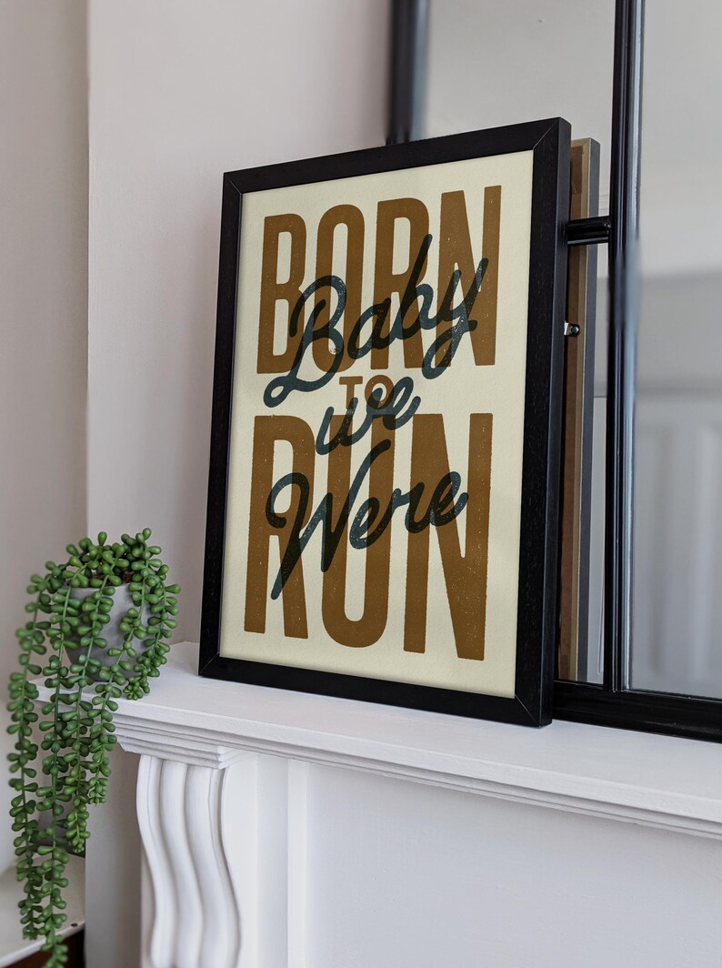 May include: A framed print with the text "Born to Run" in a brown font with the word "Baby" in a black font. The print is on a white background and is framed in black.