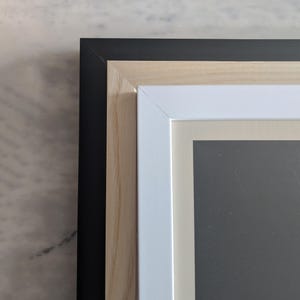 May include: Close-up of the corner of a picture frame with three layers of framing. The outer frame is black, the middle frame is natural wood, and the inner frame is white.