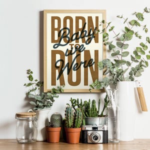May include: A framed print with the text "Baby, we were born to run" in a vintage style font. The print has a brown background with white text and is framed in a light wood frame. The print is sitting on a table with plants and other decorative items.