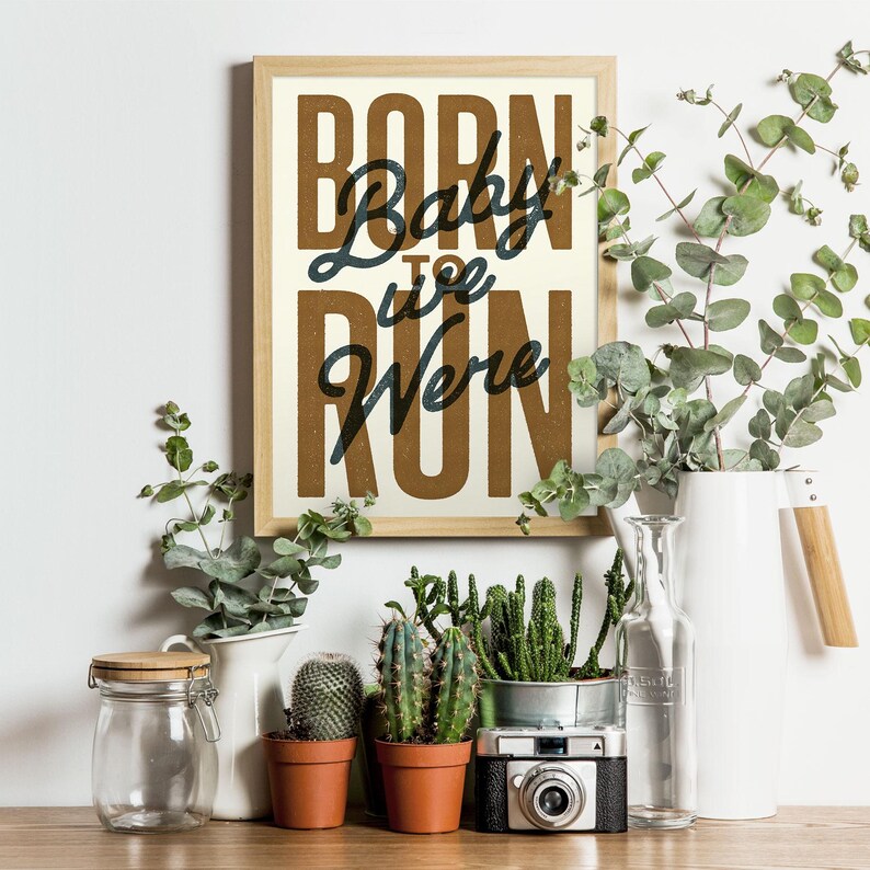 May include: A framed print with the text "Baby, we were born to run" in a vintage style font. The print has a brown background with white text and is framed in a light wood frame. The print is sitting on a table with plants and other decorative items.