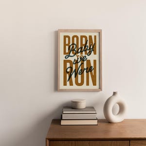 May include: A framed print with a beige background and brown text that reads "Born to Run" with the words "Baby we" in black cursive script.