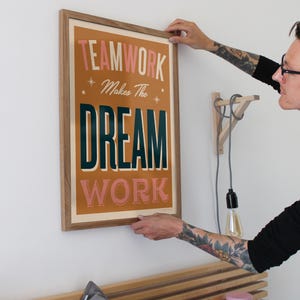 May include: A framed poster with the text "TEAMWORK Makes The DREAM WORK" in a retro style font. The poster has a brown background with pink and green text. The poster is hanging on a white wall.
