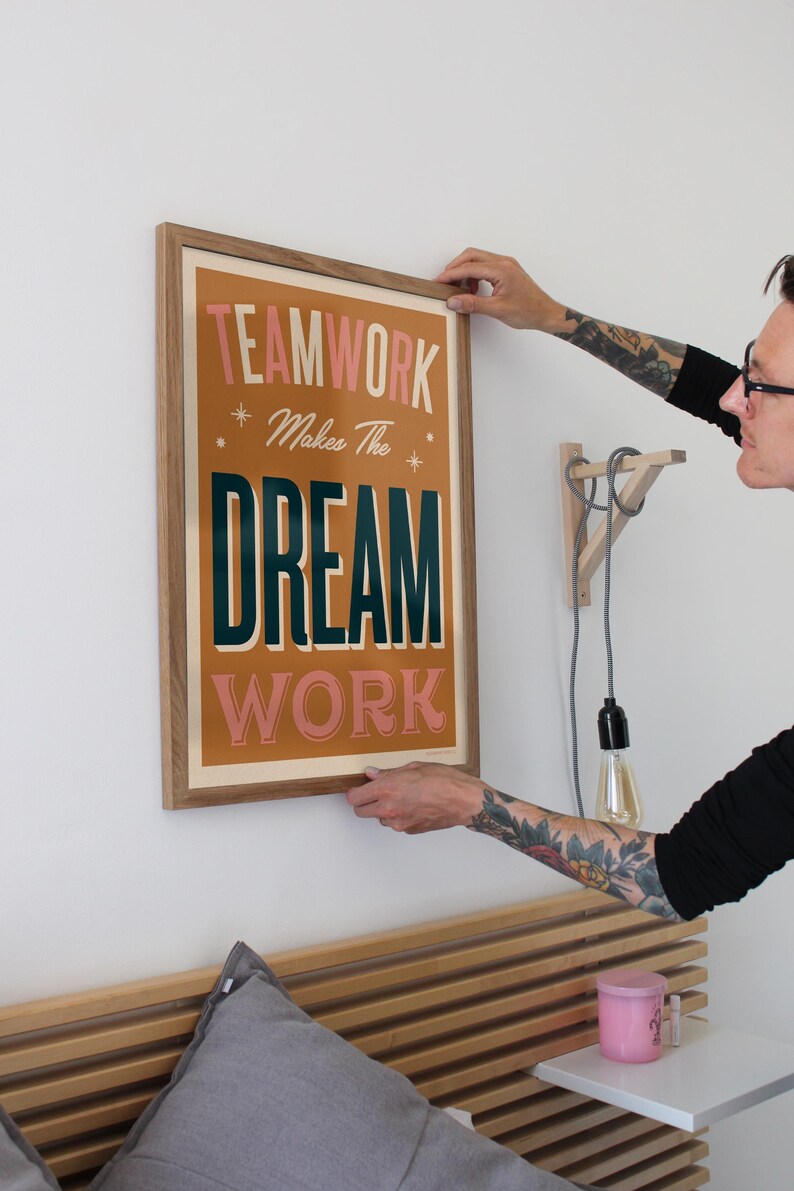 May include: A framed poster with the text "TEAMWORK Makes The DREAM WORK" in a retro style font. The poster has a brown background with pink and green text. The poster is hanging on a white wall.