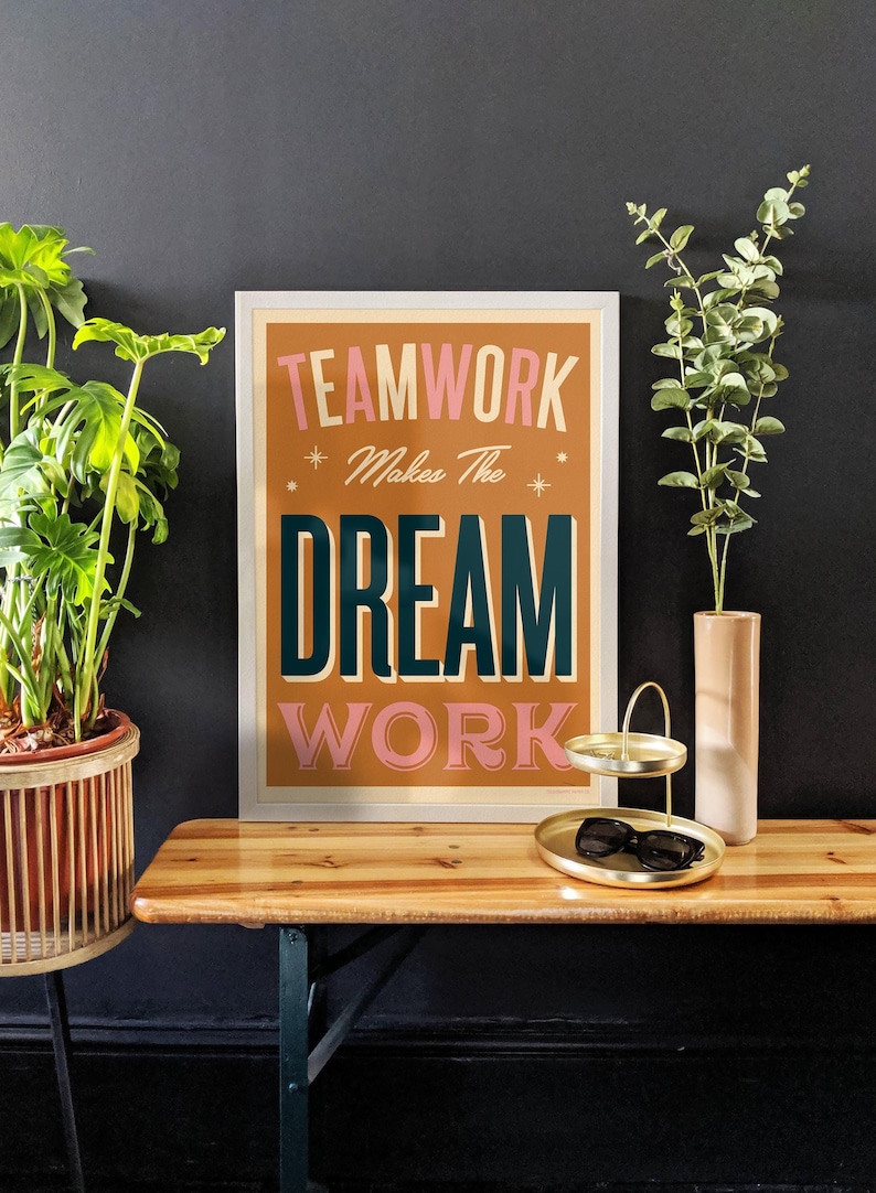 May include: A framed motivational poster with a brown background and white frame. The poster features the text "TEAMWORK Makes The DREAM WORK" in pink, white, and teal lettering.