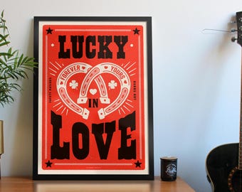 May include: A red and black poster with the text "Lucky in Love" and two intertwined horseshoes with hearts and shamrocks. The poster is framed in black.