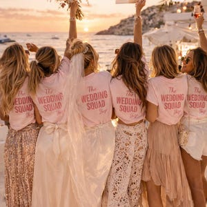 Op de afbeelding: Een groep mensen draagt roze t-shirts met "The Wedding Squad Ibiza" op de rug, met een boeket, waaier en drankjes. Ze staan op een strand bij zonsondergang en dragen verschillende rokken en shorts.