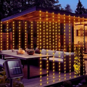 May include: An outdoor patio at dusk, lit by warm yellow string lights. A solar panel is in the foreground. The lights hang like curtains, creating a cosy ambiance around a wooden table and seating area. The scene suggests a relaxing outdoor space.